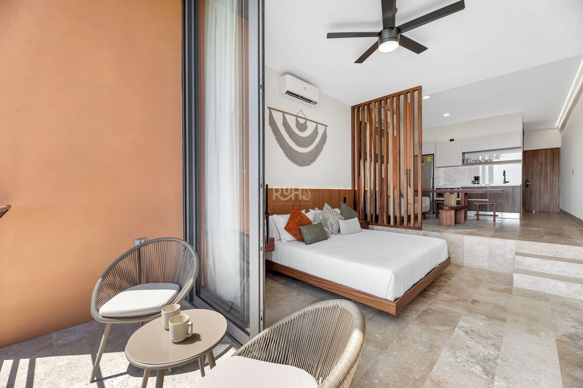Une chambre d’hôtel moderne avec un lit, deux fauteuils en osier, une petite table, un ventilateur de plafond et, au fond, une kitchenette.