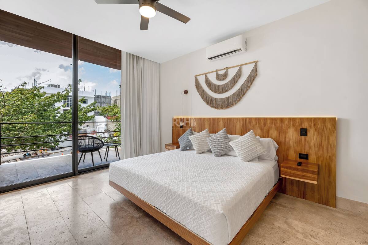 Ein modernes Schlafzimmer mit einem großen Bett, einem Holz-Kopfteil, einem Nachttisch, einer Wand-Klimaanlage und Blick ins Freie.