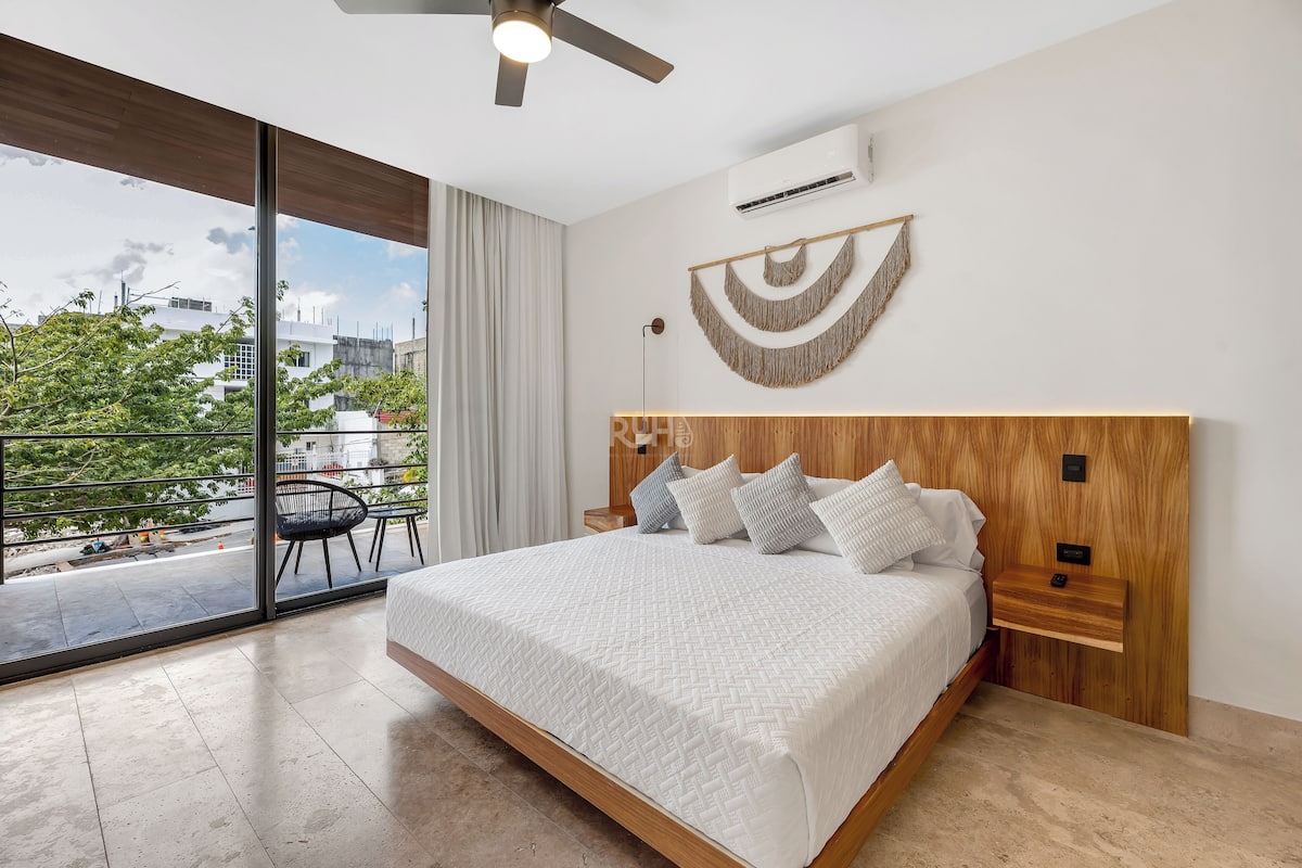A modern bedroom with a large bed, a wooden headboard, a bedside table, a wall-mounted air conditioner, and a view of the outdoors.