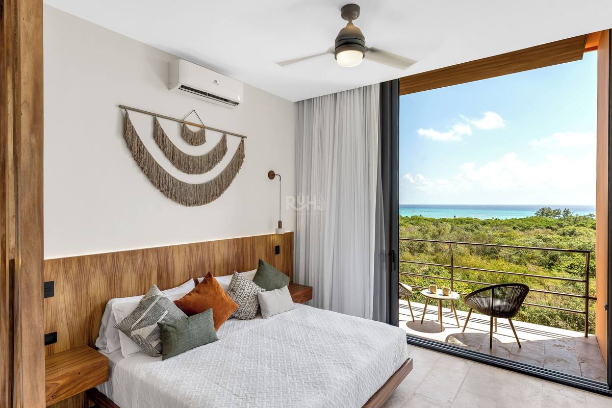 Ein Schlafzimmer mit Bett, Nachttischen, Deckenventilator, Klimaanlage, Balkon mit Aussicht und großem Fenster.