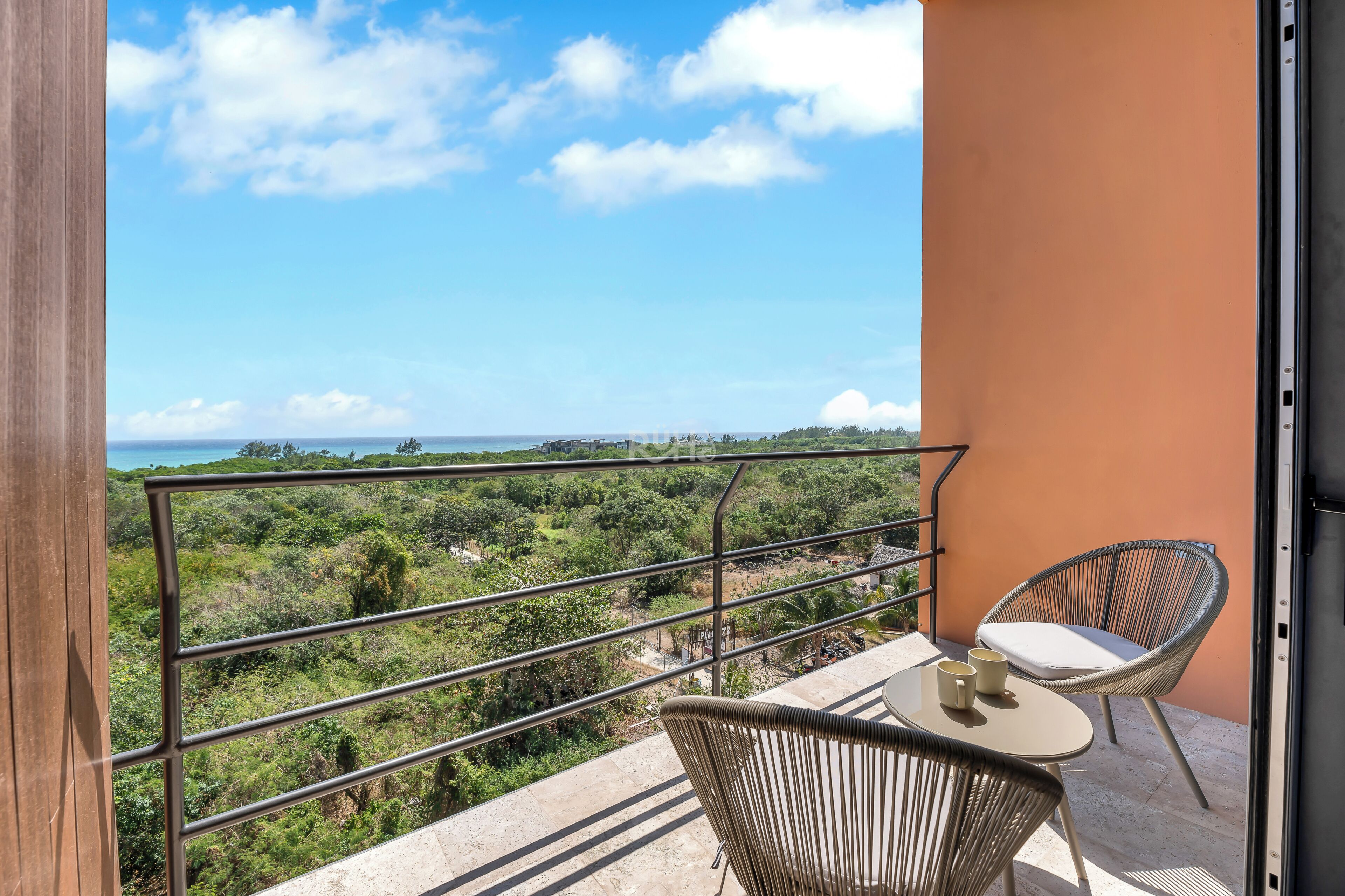 A balcony with a metal railing, two wicker chairs, a small table, and a view of a green landscape.