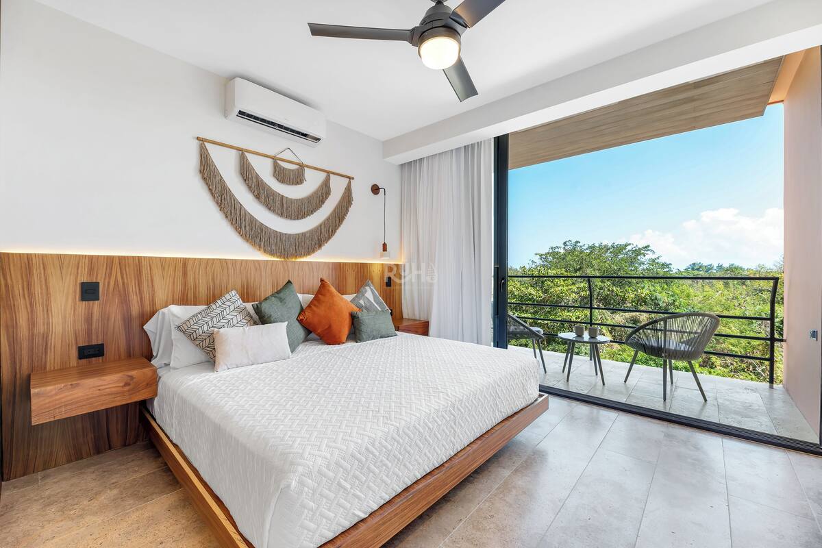 Ein modernes Schlafzimmer mit einem großen Bett, einem Deckenventilator, einer Klimaanlage, einem Holz-Kopfschild, einem Balkon mit Tisch und Stühlen sowie Blick auf Grünflächen.