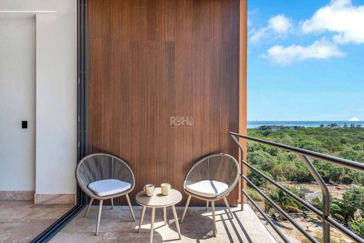 Ein Balkon mit zwei Korbstühlen, einem kleinen Tisch, einer Holzvertäfelung und Blick auf Grünflächen und das Meer.