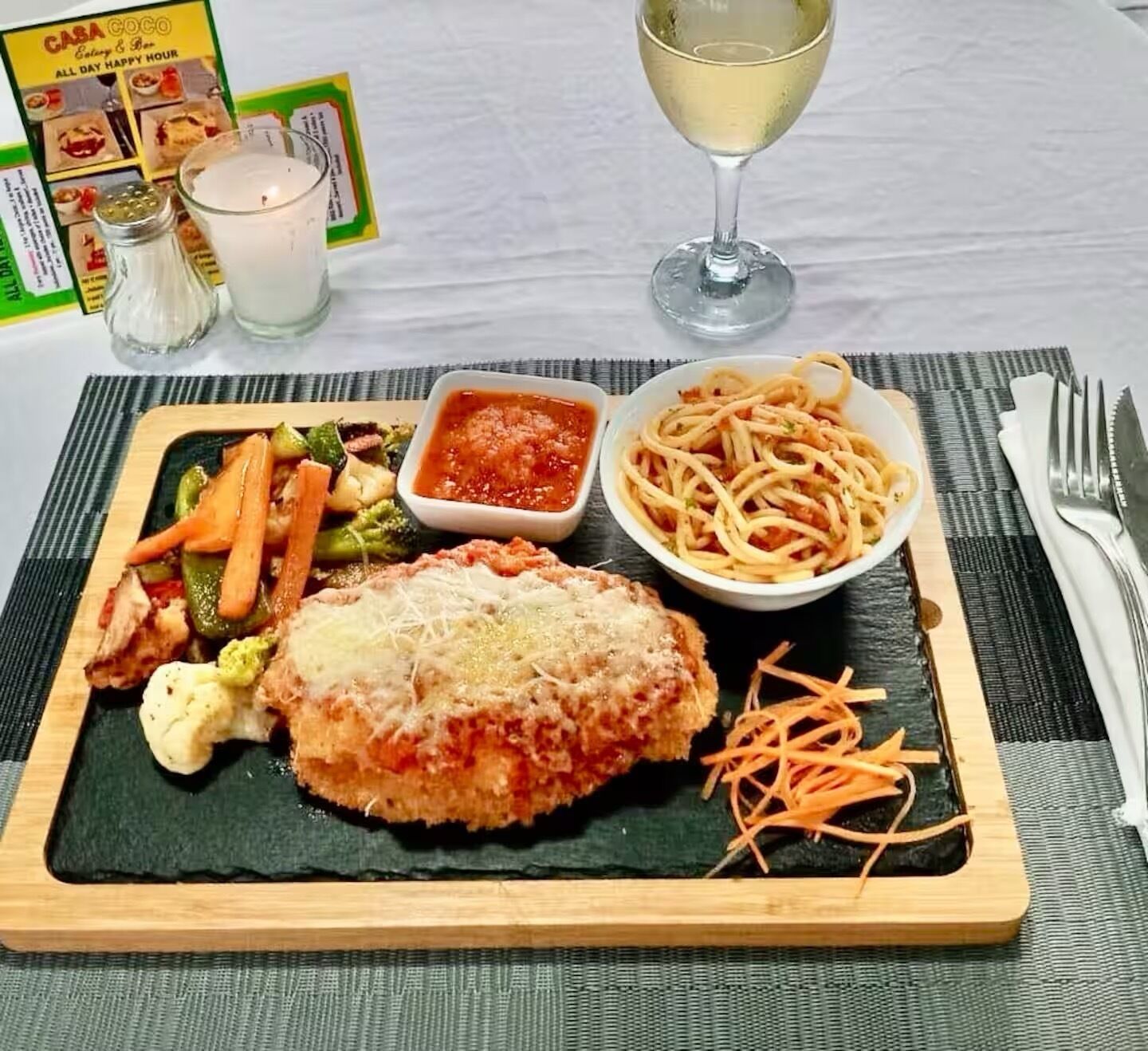 A meal with a breaded cutlet, vegetables, spaghetti, and a glass of white wine on a table.