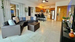 Ein modernes Wohnzimmer mit Ecksofa, Couchtisch und Essbereich mit Stühlen. Es gibt eine Küche mit Kühlschrank und Mikrowelle.