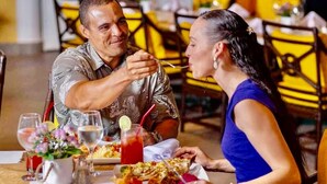 Ein Esstisch mit einem Mann und einer Frau beim Essen, einem Glas roter Flüssigkeit und einer weißen Karaffe mit Blumen.