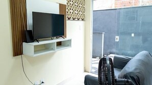 Een woonkamer met een televisie op een witte plank, een zwarte ventilator en een grijze bank.