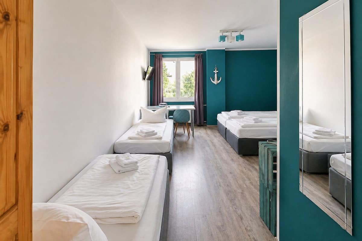 Une chambre d’hôtel avec deux lits simples, un bureau, une chaise, une fenêtre avec des rideaux et un mur d’accent de couleur turquoise.