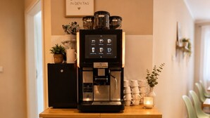 Un coin cuisine comprenant une machine à café, un meuble avec des tiroirs, un panneau décoratif accroché au mur et une plante posée sur le comptoir.