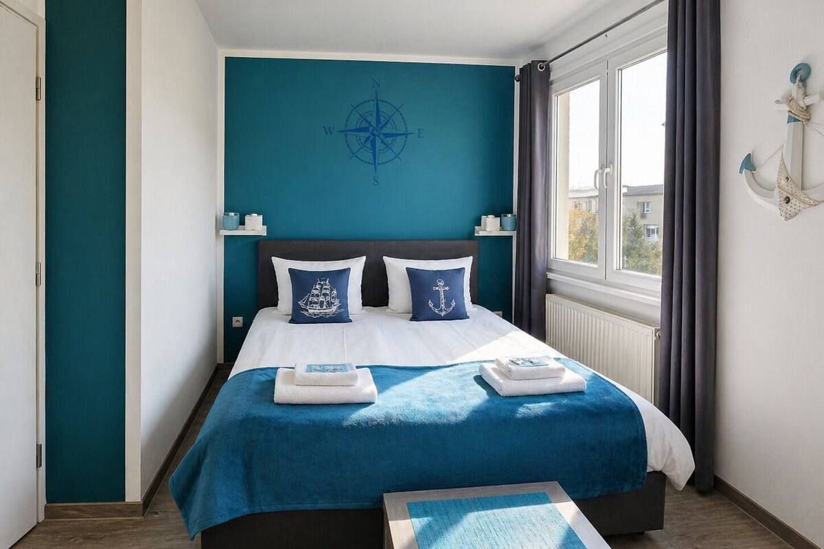 Une chambre avec un lit, une fenêtre, un décor mural représentant une boussole et une petite table.