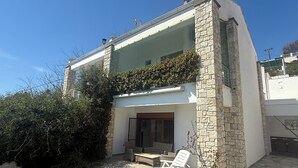 Une maison moderne avec une façade en pierre, un balcon en verre et une terrasse aménagée avec du mobilier d’extérieur.