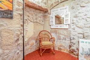 A wicker chair in a stone-walled room with a red floor, a white decorative panel, and a poster on the wall. - Charming 1750 Stonehouse in the heart of Saint Rémy (Saint-Rémy-de-Provence)