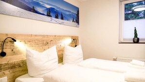 A hotel room with a wooden headboard, two white beds, a nightstand, a wall-mounted lamp, a window with blinds, and a painting of a snowy landscape. - Helles und Modernes Appartement (Winterberg)