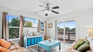 A living room with a sofa, a coffee table, a ceiling fan, a cabinet, and a large window with a view. - Beachfront Duplex 6BR/4BA – 16+ Guests – Paradise Park, Melbourne FL (Melbourne)