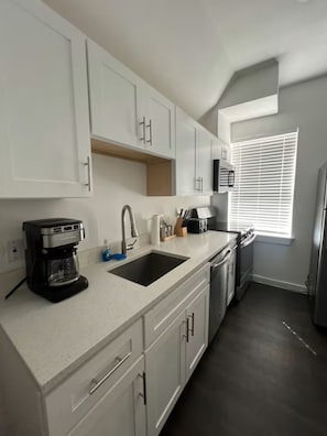 A modern kitchen with white cabinets, a coffee maker, a sink, and a window with blinds. - Sweet W. Campus Getaway - Rest & Relax for SXSW (Austin)