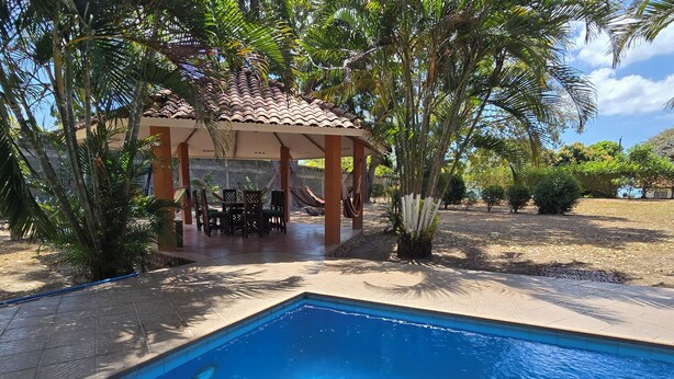 Una piscina con solárium de baldosas, un área de estar al aire libre techada con sillas y mesas, y palmeras circundantes.