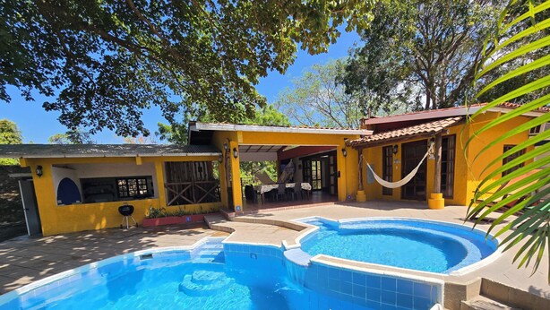 Una piscina con borde de azulejos, un patio cubierto y un edificio amarillo con techo rojo.