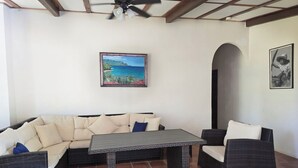 Una habitación con un sofá, dos sillas, una mesa, un ventilador de techo y un cuadro que representa una playa.