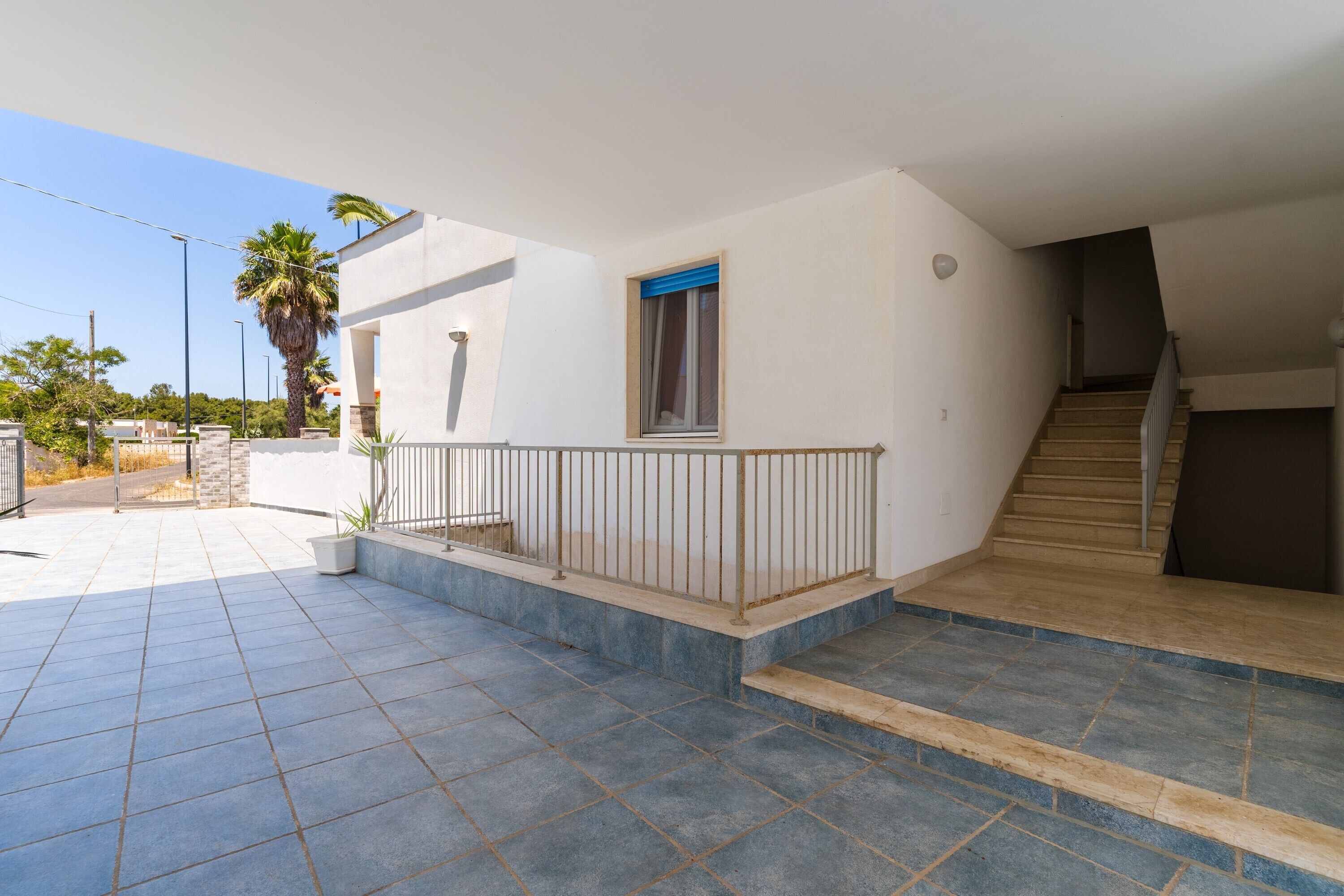 A residential building with a tiled patio, a small window with blue curtains, and a staircase leading to an upper floor.