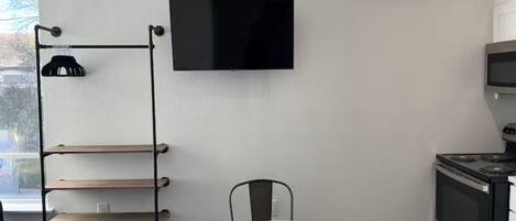 Une cuisine moderne dotée d’un four encastré, d’une table à manger avec des chaises, d’un téléviseur fixé au mur et d’une étagère métallique à plusieurs niveaux.