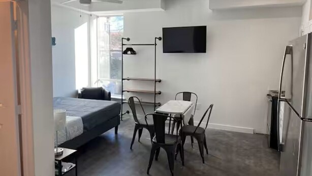 A modern room with a bed, a dining table with chairs, a TV mounted on the wall, and a refrigerator. - ATX Nice Studio Walking distance to the Drag! (Austin)