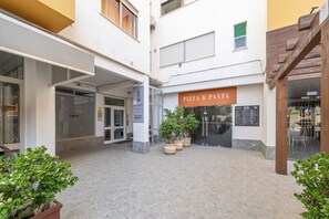 Ein Gebäude mit einem 'PIZZA & PASTA'-Schild, einem Glastür-Eingang und Kübelpflanzen im Innenhof.