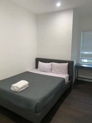 A single bed with a grey mattress and pillows, a small bedside table, and a window with blinds. - Spacious 6x3 Apt near UT (Austin)