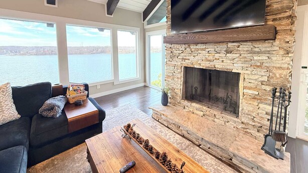 A living room with a stone fireplace, a flat-screen TV, and a large window overlooking a body of water. - Wide Sky Chateau Lake Views (Osage Beach)