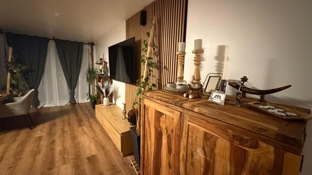 Un salon avec un plancher en bois, un téléviseur fixé au mur, un meuble en bois orné d’objets décoratifs et une plante.