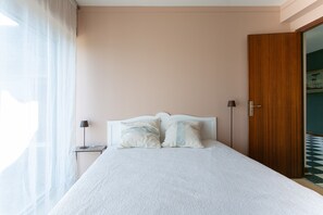 Un dormitorio con cama, mesitas de noche, lámpara, una puerta y vistas a través de una ventana.