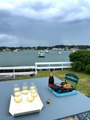 Una mesa con copas de vino, una botella y alimentos, con vista a un cuerpo de agua y barcos.