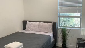 Ein Schlafzimmer mit einem Bett, einem Fenster mit Jalousien, einer Zimmerpflanze und einem Nachttisch.