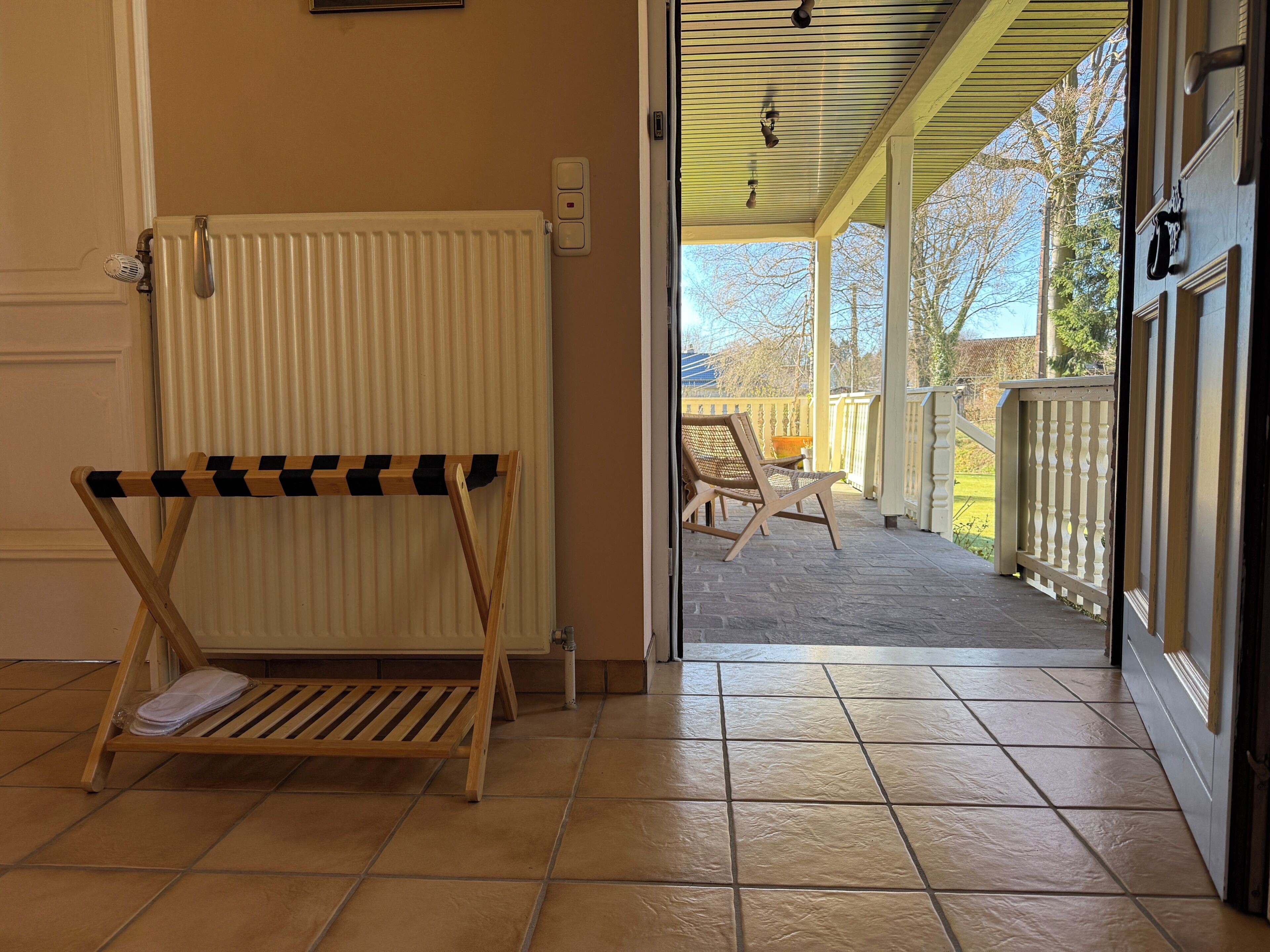 Una habitación con un radiador, un banco de madera y vistas a un patio exterior con sillas y una barandilla.