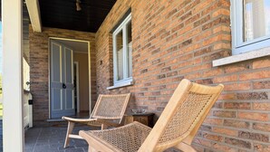 Twee rieten loungestoelen op een terras van bakstenen, met een deur en raam op de achtergrond.