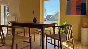 Een eetruimte met een houten tafel en stoelen, een vaas met een plant, en uitzicht op buiten.