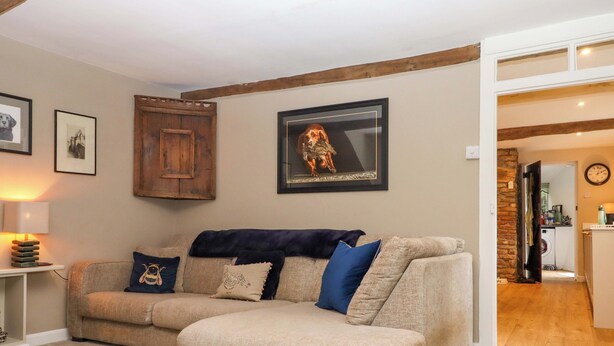 Een woonkamer met een bank, ingelijste foto’s, een lamp en een keuken die zichtbaar is door een open deur.