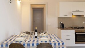 A dining area with a checkered tablecloth, a bottle of wine, and a kitchen with white cabinets and a stove. - Apartment I Pini by Interhome (Bibbona)