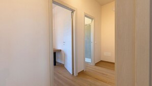 Un corridoio con pavimento in legno, pareti bianche e una porta che dà su un bagno con box doccia in vetro.