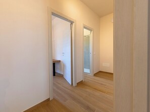 A hallway with wooden flooring, white walls, and a door leading to a bathroom with a glass shower. - Apartment I Pini by Interhome (Bibbona)