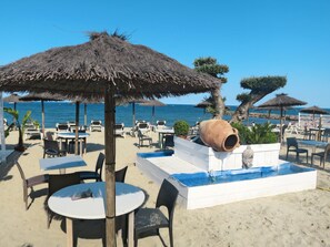 Ein Sitzbereich im Freien mit Tischen und Stühlen, ein großer Terrakottatopf und ein kleines Wasserspiel am Sandstrand mit Sonnenschirmen und Palmen.