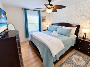 A bedroom with a bed, a ceiling fan, a nightstand, a lamp, a dresser, a window with curtains, and a wall with a textured pattern. - 🌴🍹Rustic Coastal Retreat with Patio, BBQ, & Privacy Fenced Yard! Near Beaches! (Sebastian)