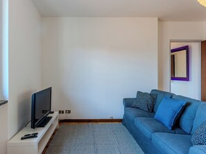 Ett vardagsrum med en platt-TV på ett vitt TV-bord, en blå soffa med mönstrade kuddar och en grå matta.