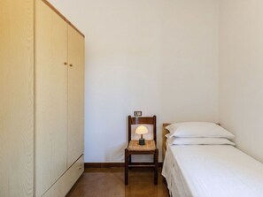 Ein kleines Zimmer mit einem Einzelbett, einem Holzhocker mit Lampe und einem Kleiderschrank.