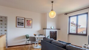 A living room with a sofa, a dining table with chairs, a hanging light fixture, and a window with a view of buildings. - Casa Pradell by Interhome (Gravedona)