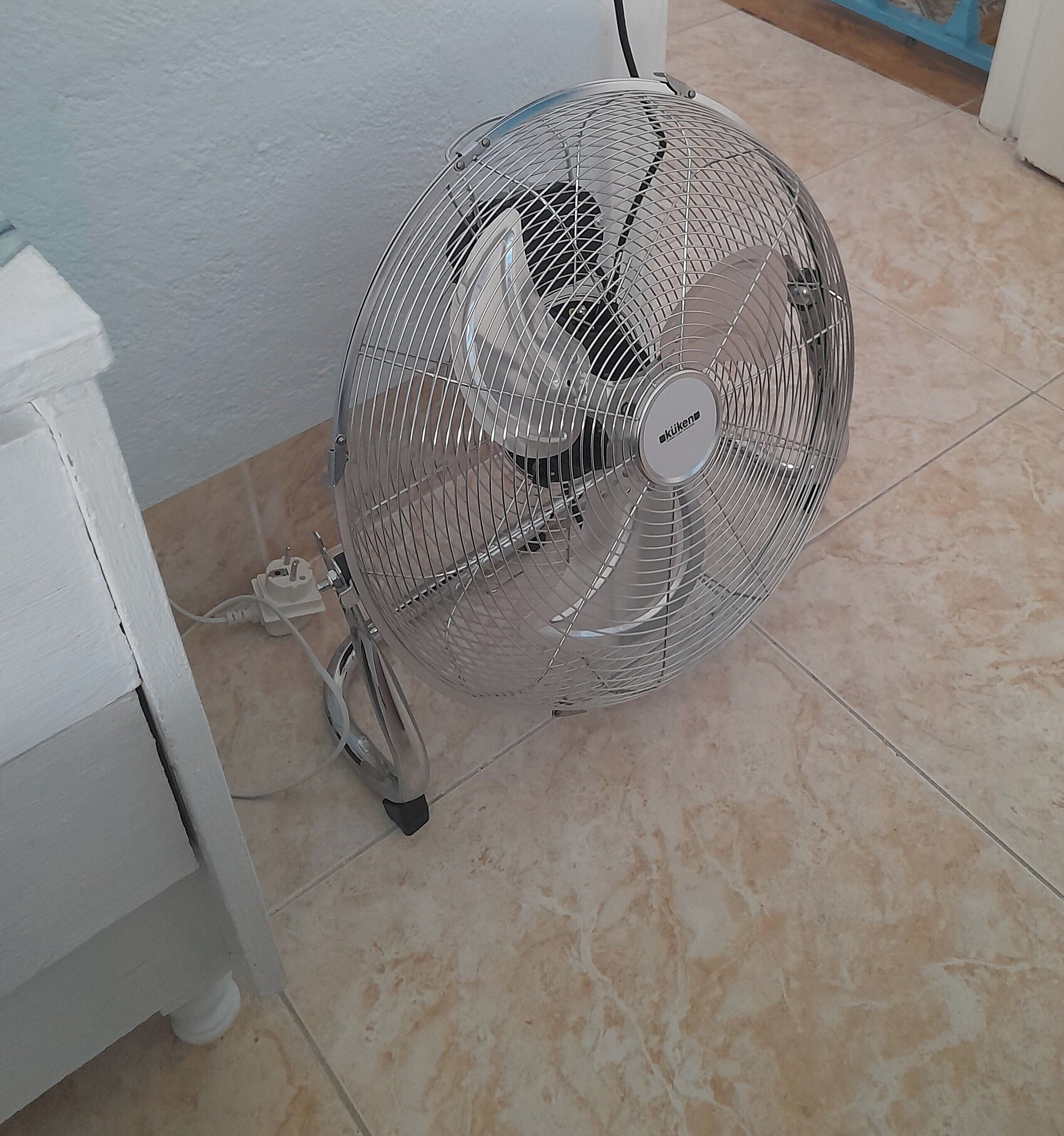 Un ventilador de pie con rejilla metálica y cable de alimentación conectado, sobre un suelo de baldosas junto a una pared blanca.