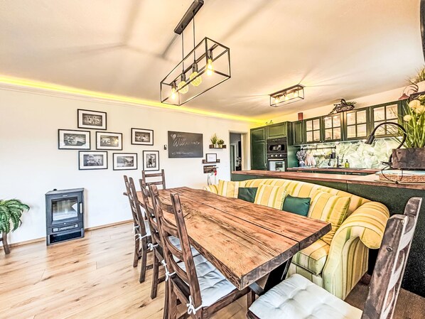 A dining area with a wooden table and chairs, a bench, a kitchen with green cabinets, and framed pictures on the wall.