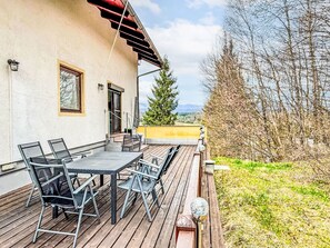 A wooden deck with a table and chairs, a window, and a view of trees and mountains.