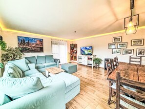 A living room with a sofa, a TV, a dining table with chairs, and a wall with framed pictures.