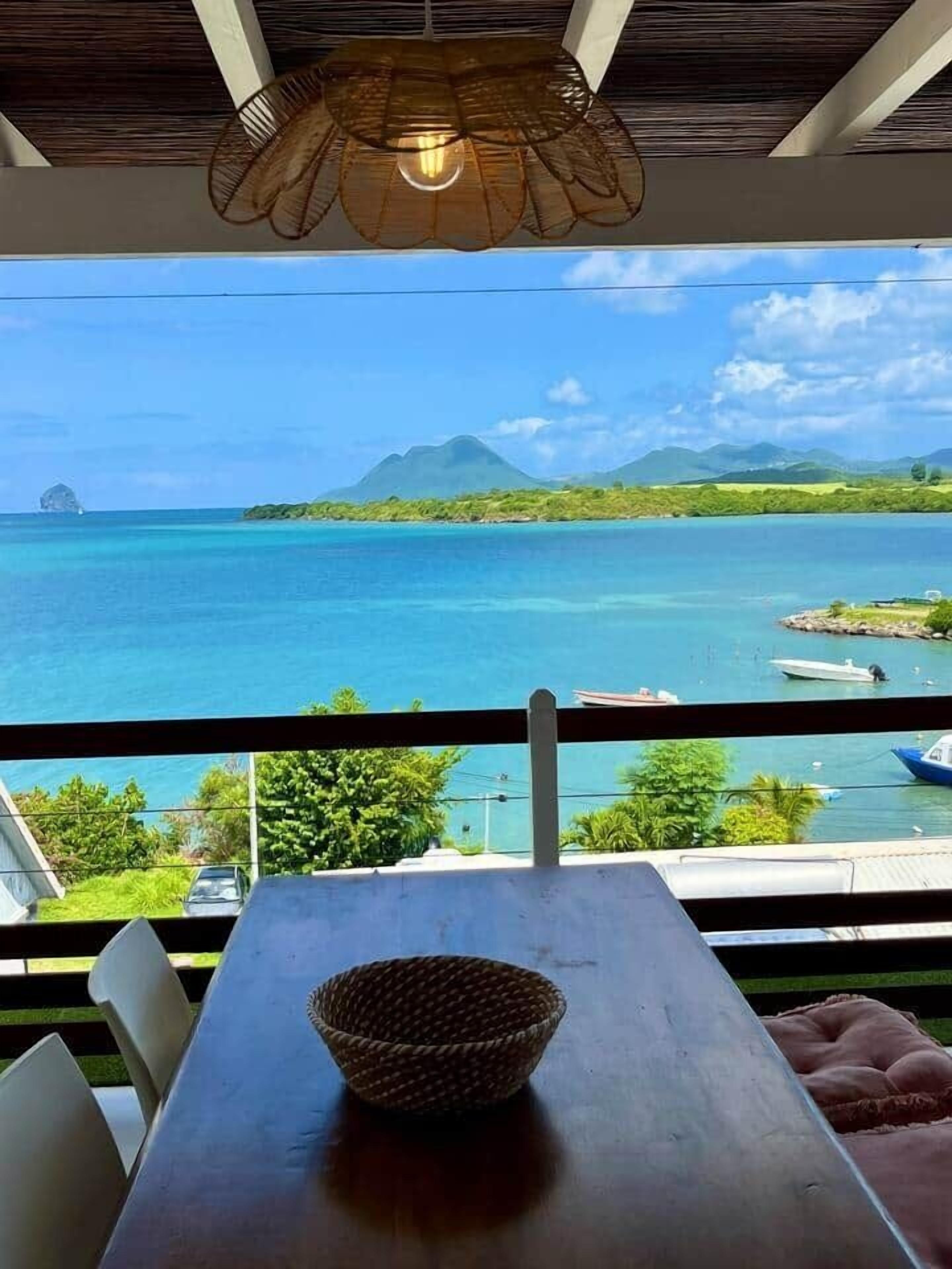 Un balcó amb vista al mar turquesa, una taula de fusta i una cistella teixida, juntament amb una làmpada penjant.