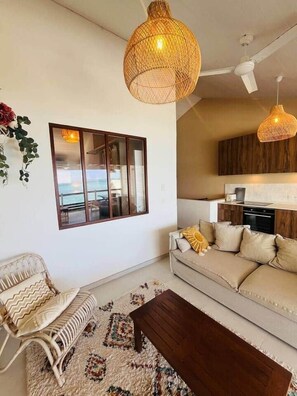 A living room with a sofa, a coffee table, a wicker pendant light, a wall-mounted picture, and a view through a glass door. - Apartment KayTi palm trees (Sainte-Luce)